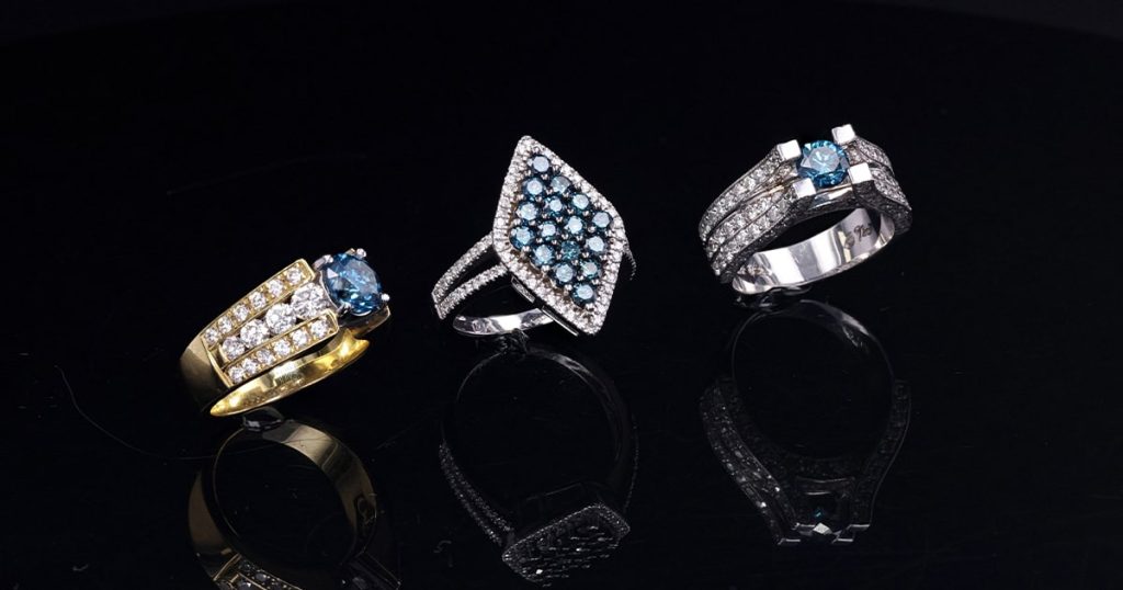 Three unique blue diamond rings displayed on a reflective black surface — one in yellow gold with diamond accents, one marquise-shaped cluster design in white gold, and one modern white gold band with a solitaire blue diamond and pave diamonds.