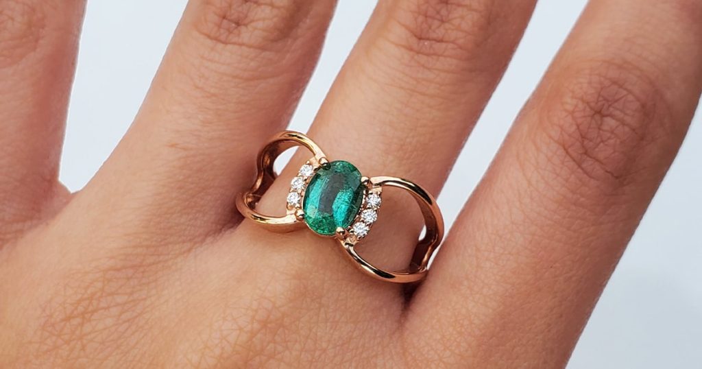 Close-up of a hand wearing a rose gold ring featuring an oval green emerald accented with small diamonds on each side.