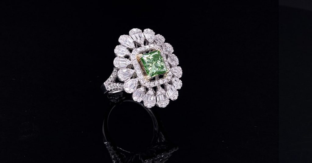 White gold ring with a square-cut green diamond center stone, surrounded by a double halo and layered cluster of baguette and round diamonds, displayed on a black background.