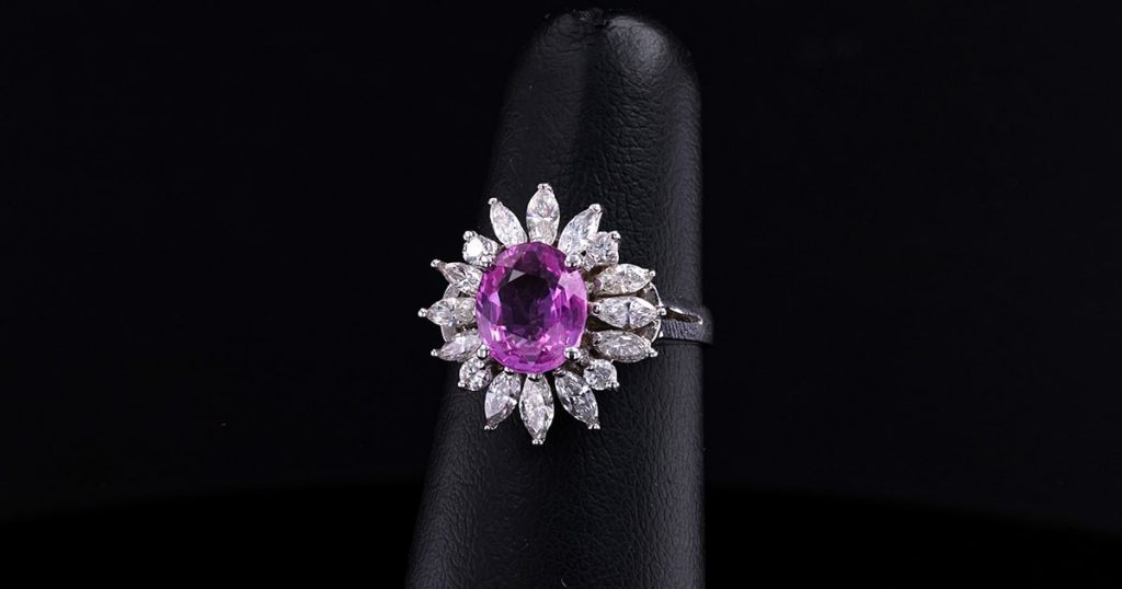 Oval pink sapphire ring with a diamond sunburst halo design set in white gold, displayed against a black background.