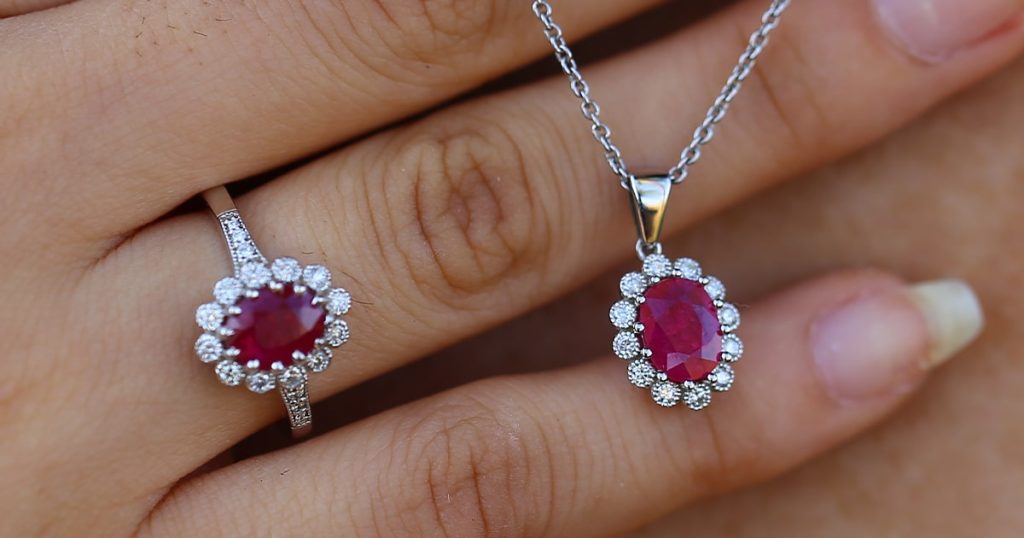 Close-up of a hand wearing a ruby and diamond halo ring, paired with a matching ruby and diamond halo pendant on a silver chain.