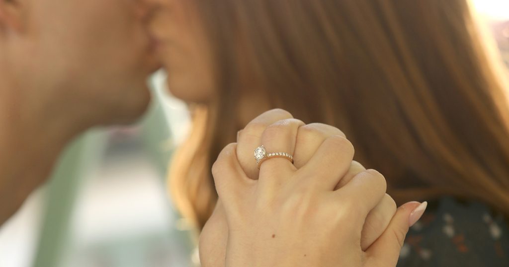 Couple holding hands while kissing, with a diamond engagement ring on her finger in focus.