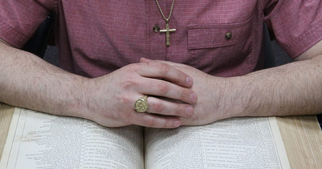 Man wearing a gold cross necklace and ring, hands clasped over an open Bible