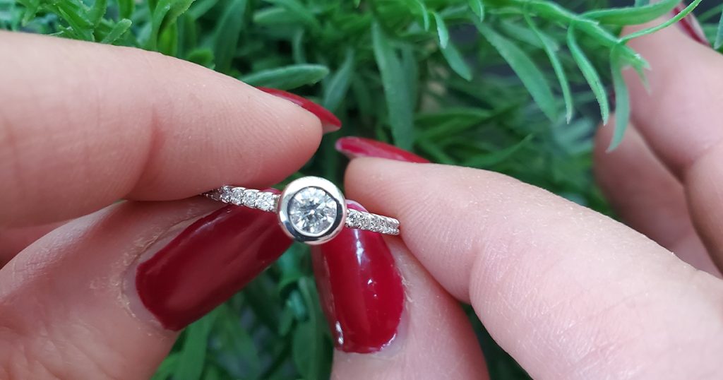 Close-up of a bezel-set diamond engagement ring with pavé band, held between hands with red polished nails.