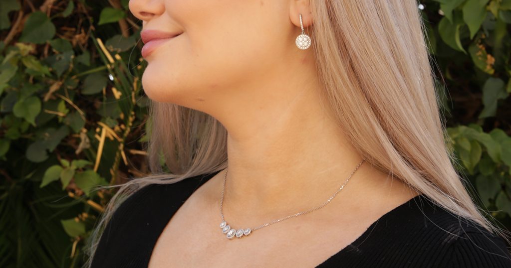 Close-up of a woman wearing diamond drop earrings and a diamond necklace with five sparkling stones, against a natural outdoor background.