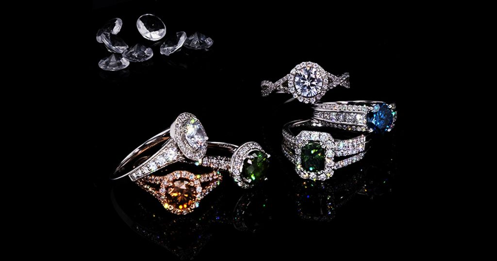 Collection of diamond halo rings with different center stones — white, green, blue, and cognac — displayed on a reflective black surface with loose diamonds in the background.