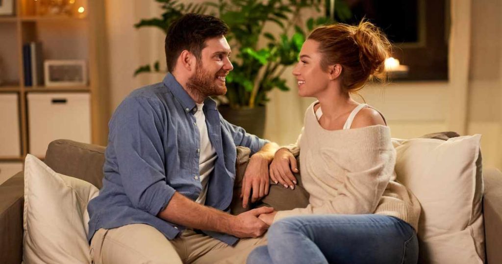 A happy couple sitting on a cozy couch at home, smiling warmly at each other while holding hands, with soft lighting and plants in the background.