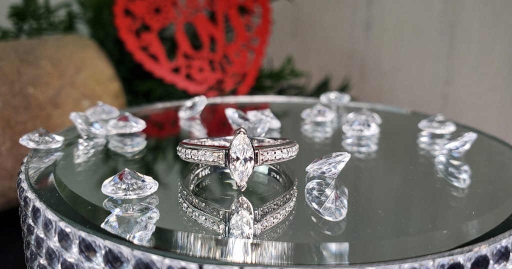 Marquise-cut diamond engagement ring with pavé band, displayed on a mirrored surface surrounded by loose diamonds.