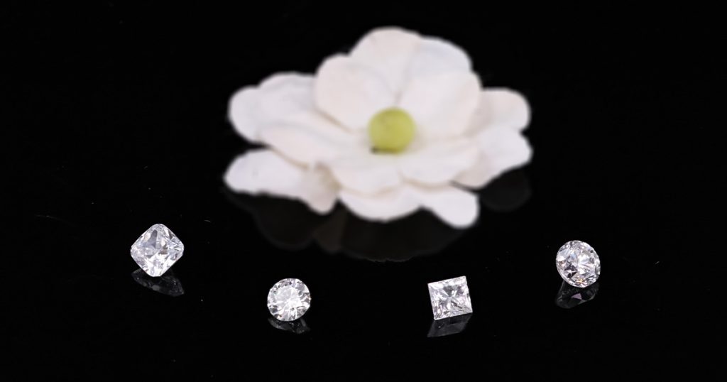 Four loose diamonds in different cuts — cushion, round, princess, and oval — displayed on a reflective black surface with a white flower in the background.